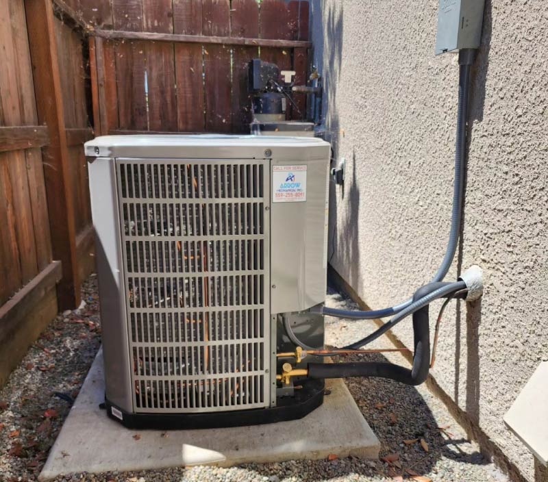 A newly installed outdoor AC heat pump unit with Arrow HVAC contact sticker, positioned on a concrete pad next to a stucco house wall and wooden fence in Fowler.