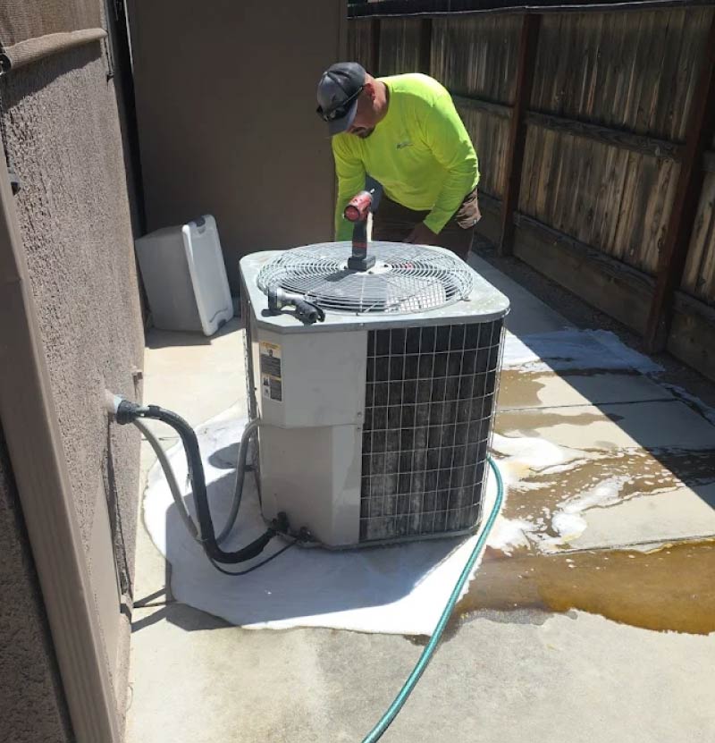 An Arrow HVAC technician cleaning and servicing a residential outdoor air conditioner condenser unit with a pressure hose, performing ac repair in selma, ca