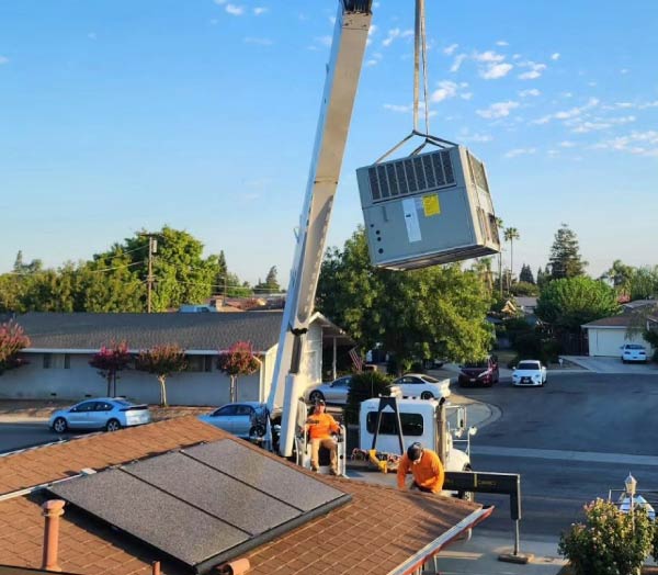 A crane hoisting a large commercial or residential rooftop AC unit during an HVAC replacement or installation in a neighborhood.