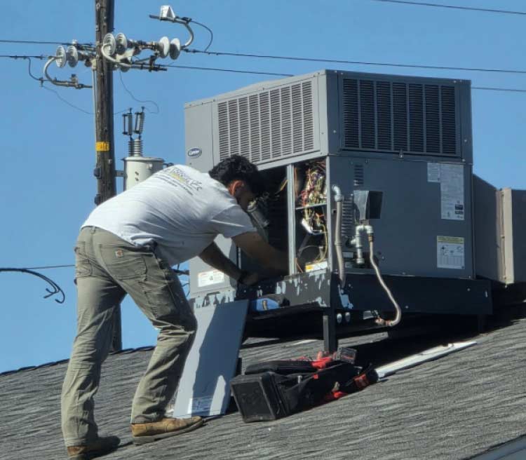 HVAC service technician on a shingled roof repairing a large commercial Carrier air conditioning unit with its side panel open, showing wiring.