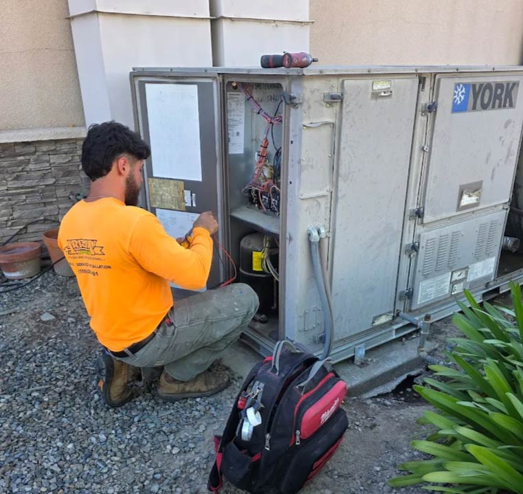 An Arrow HVAC technician in an orange long-sleeve shirt kneeling to repair and diagnose a large outdoor commercial York AC unit on a gravel surface, for ac repair in fresno, ca