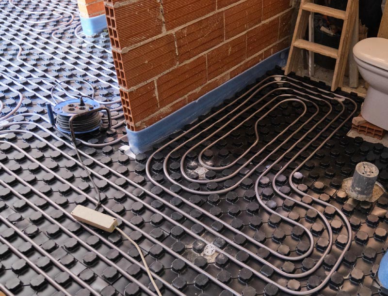Underfloor radiant heating system installation in a bathroom under renovation, featuring flexible piping snaked through black knobbed insulation trays near an exposed brick wall and a toilet base.
