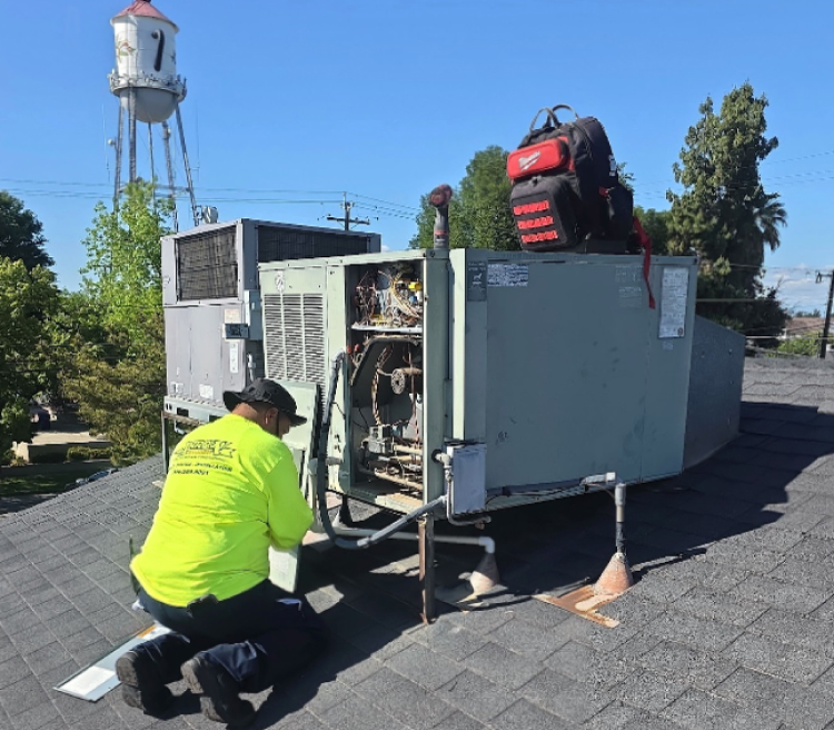 This image shows an HVAC technician wearing a bright, high-visibility neon yellow shirt, dark pants, and a sun hat, kneeling on a shingled residential roof. He is performing service or diagnostics on a large, grey packaged HVAC rooftop unit. An access panel has been removed from the side of the system, exposing internal electrical wiring, components, and circuitry. A red and black Milwaukee tool backpack rests on top of the main unit. In the background, another HVAC unit is visible, along with green trees and a prominent white water tower under a clear, bright blue sky.