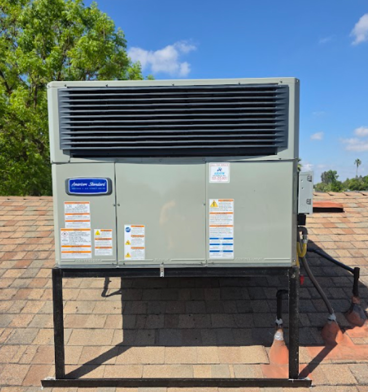 This image features a close-up, front-facing view of a clean, light grey American Standard packaged HVAC unit neatly installed on a sloped roof with brown and tan shingles. The unit is mounted on a sturdy black metal support frame to keep it level. It features a large black horizontal grille at the top, a prominent blue "American Standard" brand badge on the left panel, and multiple safety and informational warning labels affixed to the front panels. The background shows a bright blue sky with light, scattered clouds and the tops of green trees.