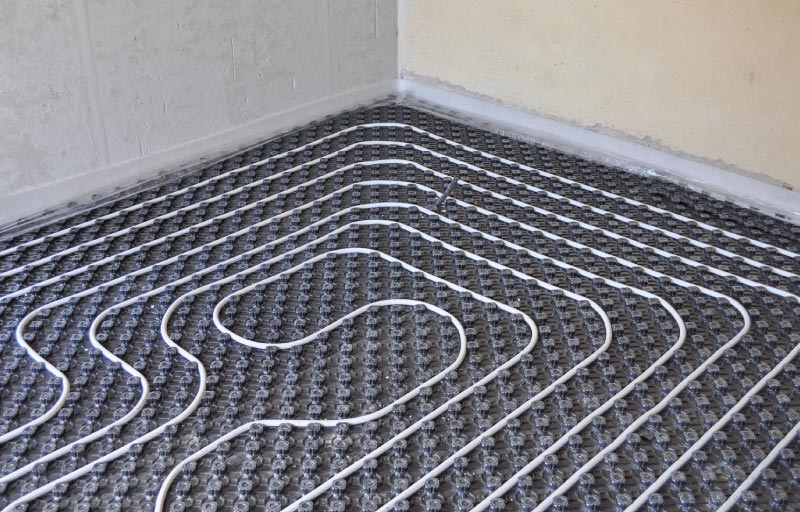 : A wide shot of an empty room under construction showing white radiant heating pipes laid out in a systematic serpentine loop across a grey textured underlayment.
