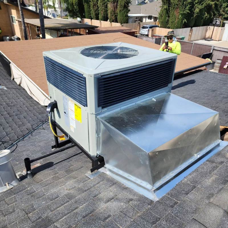 A completed packaged rooftop air conditioner installation showcasing shiny new custom sheet metal ductwork on a shingled roof with an HVAC technician standing in the background.