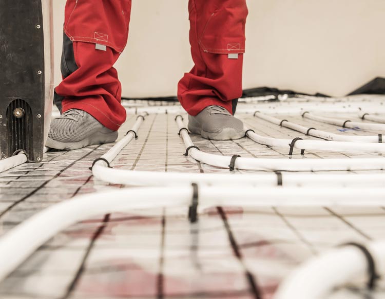 Close-up of an HVAC technician's legs in red work pants and grey shoes standing on a reflective subfloor grid while installing white underfloor radiant heating tubes.