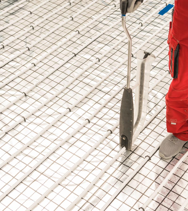 High-angle view of a worker using an installation tool to secure parallel rows of white radiant heating tubes onto a silver, gridded insulation mat.