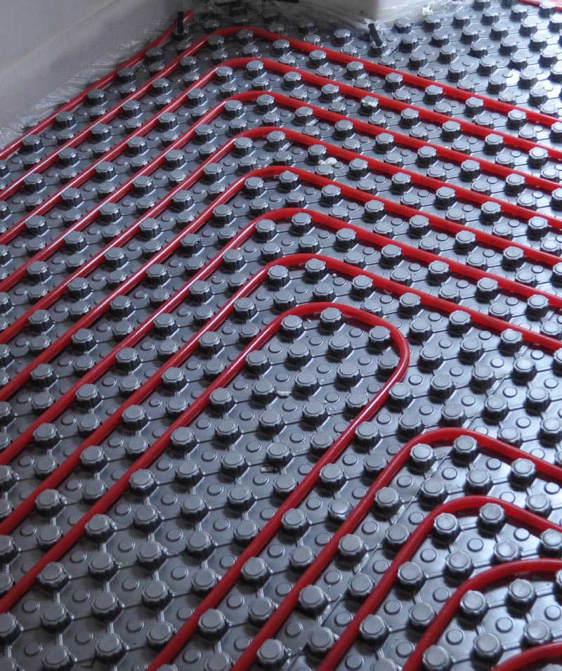 Overhead view of bright red PEX piping laid out in a continuous winding pattern across a grey, knobbed radiant heating repair floor insulation panel.