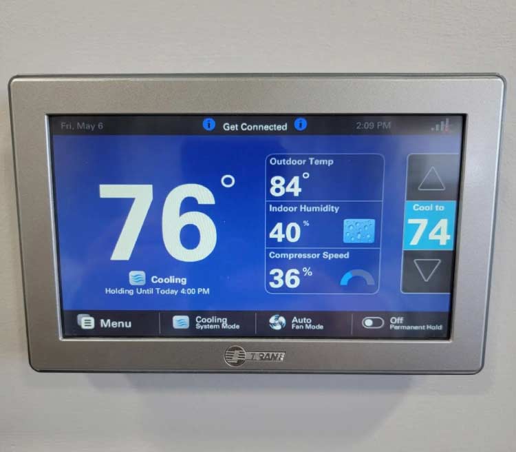 Trane smart touchscreen thermostat mounted on a wall displaying an indoor temperature of 76 degrees, outdoor temperature of 84 degrees, and cooling target set to 74 degrees.
