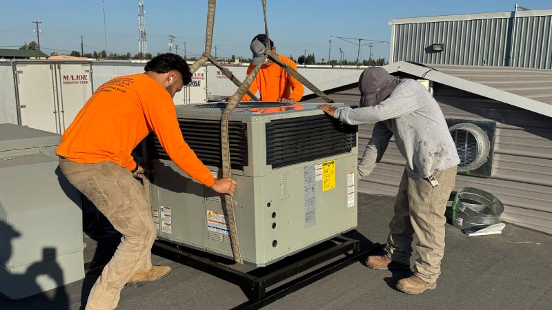 HVAC technicians wearing orange work shirts installing a large commercial rooftop AC unit using crane straps.