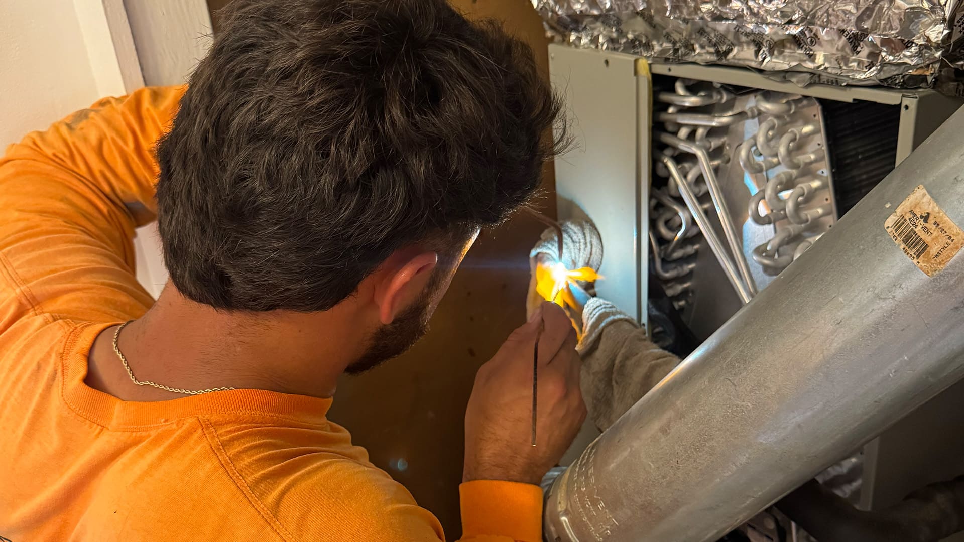 An Arrow HVAC technician in an orange shirt using a torch to repair an indoor evaporator coil system.