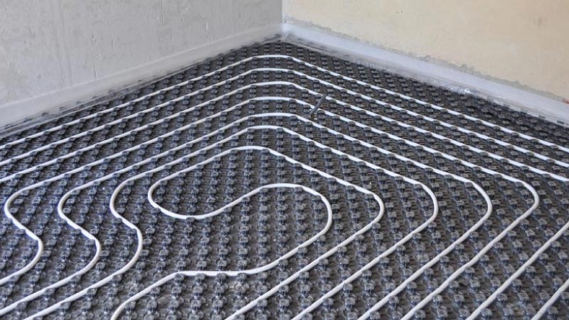 A wide shot of an empty room under construction showing white radiant heating pipes laid out in a systematic serpentine loop across a grey textured underlayment.