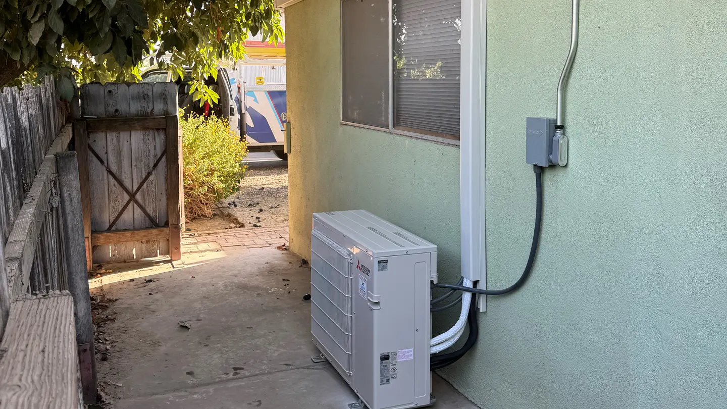 Side view of an outdoor mini-split condenser unit connected to line set covers and an electrical disconnect box on a house exterior.