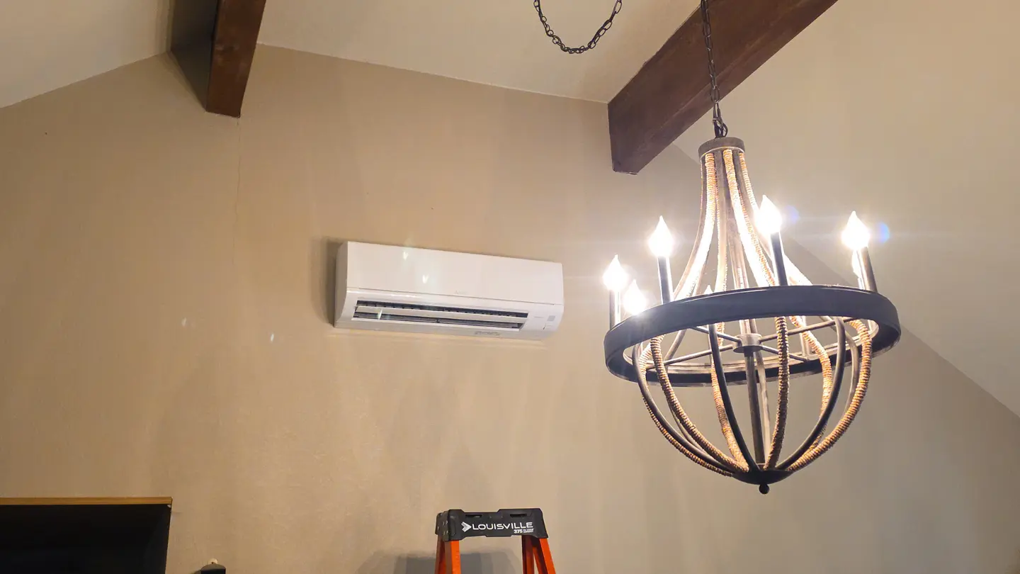 Wall-mounted indoor ductless mini-split unit installed in a room with vaulted ceilings and a rustic hanging chandelier.
